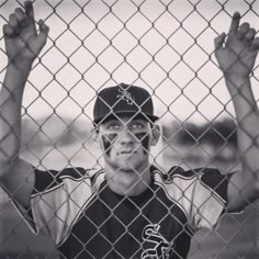 a baseball player behind a fence holding his hands up in the air and looking at the camera