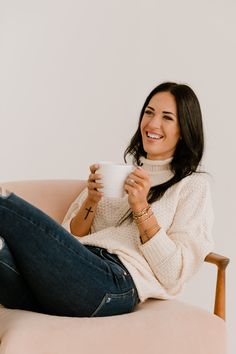 a woman sitting in a chair holding a cup and smiling at the camera while wearing jeans