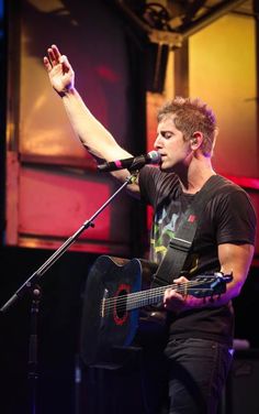 a man holding a guitar on stage with his right hand in the air and one arm raised