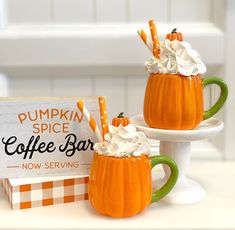 two pumpkin mugs with whipped cream and orange striped straws on them, sitting next to a sign that says pumpkin spice coffee bar now serving