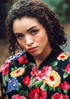 a young woman with curly hair wearing a floral shirt looking at the camera while standing outside