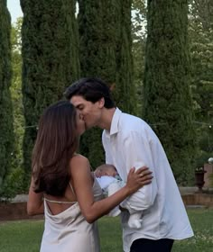 a man and woman kissing while holding a baby in front of some tall green trees