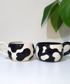 two black and white coffee mugs sitting next to each other on a table with a plant in the background