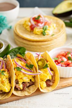 three breakfast tacos on a cutting board with salsa and avocado in the background