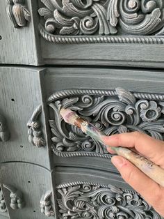 a person holding a paintbrush in front of an ornate gray dresser with carvings on it