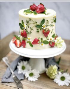 a white cake with strawberries and daisies on it sitting on top of a wooden table