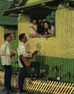 two men are playing guitar in front of a house with people on the outside wall