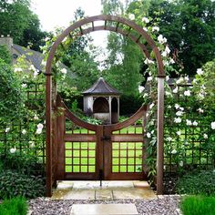 a wooden gate surrounded by white flowers and greenery with a gazebo in the background