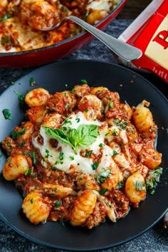 a black plate topped with pasta and meat covered in sauce next to a red pan