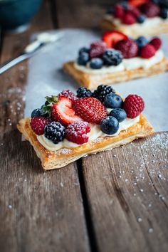 two pieces of bread with berries and cream on top sitting on a wooden table next to a bowl of strawberries