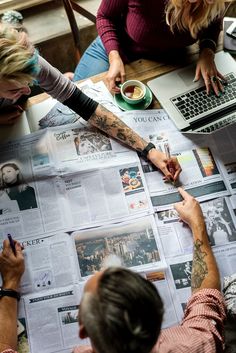 three people sitting at a table with newspapers and laptops in front of them, one person writing on a piece of paper