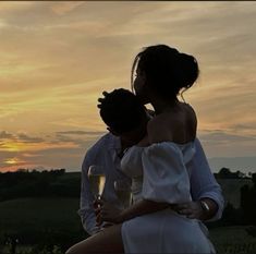 a man and woman sitting next to each other holding wine glasses in front of a sunset