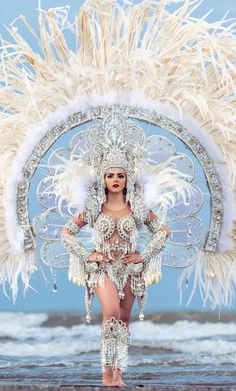 a woman in a white costume with feathers on her head and body, standing on the beach