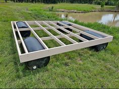 an empty bed frame sitting in the grass near a pond with water and trees behind it