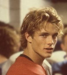 a young man in a red shirt is looking at the camera with other people behind him