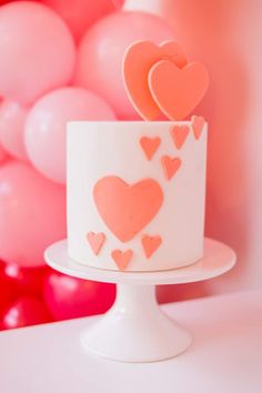 a white cake with pink hearts on top and balloons in the background at a birthday party