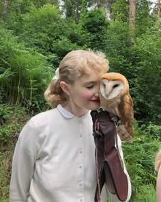 a woman holding an owl on her arm in front of trees and bushes while wearing gloves