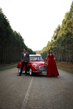 a man and woman standing next to an old fashioned red car on a road with trees in the background