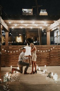 a man kneeling down next to a woman in front of a building with candles on the ground