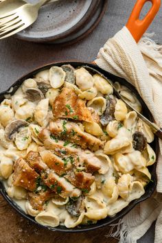 a skillet filled with pasta and meat on top of a wooden table next to utensils