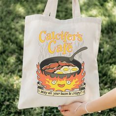 "Our Howl's Moving Castle Inspired Calcifer's Cafe 100% cotton bag comes in one size - 15\" x 16\"- perfect for everyday wear. While the canvas material will show off your designs in great colors, it's durable and will last for years. The bag features 20\" handles (made from the same canvas), making it easy to carry even with a week's worth of shopping in Ghibli inspired style! .: 100% cotton canvas .: Heavy fabric (12 oz/yd² (406.9 g/m .: Sewn-in label" Howls Moving Castle Designs, Howls Moving Castle Items, Howl's Moving Castle Embroidery, Howls Moving Castle Merchandise, Ghibli Tote Bag, Decorated Tote Bags, Eco Bags, Best Tote Bags