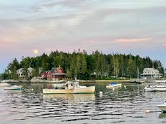 several boats floating on the water in front of some houses and trees with a full moon behind them