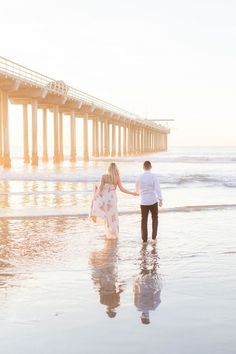 a man and woman holding hands while walking on the beach with a pier in the background