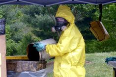 a man in a yellow coverall and gas mask is pouring out some liquid from a barrel