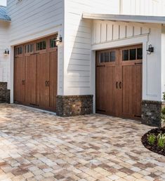 two brown garage doors on the side of a white house next to a brick driveway