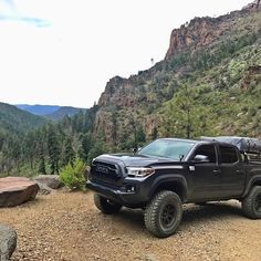 a black truck is parked on the side of a mountain with trees and mountains in the background