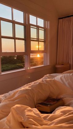 a book is laying on the bed in front of two windows with sunsets outside