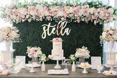 a table topped with a white cake and lots of pink flowers next to a tall green wall