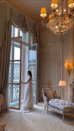 a woman standing in front of a window next to a couch and chandelier