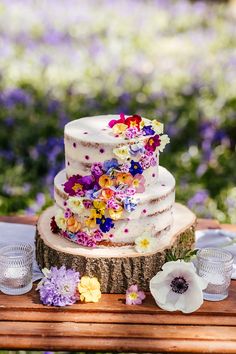 a multi - tiered wedding cake with colorful flowers on the top sits on a tree stump