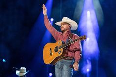 a man in a cowboy hat holding a guitar on stage with his right hand up