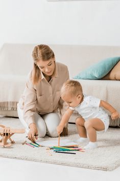 a woman kneeling down next to a baby playing with crayons on the floor