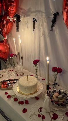 a white cake sitting on top of a table covered in red balloons and roses next to candles