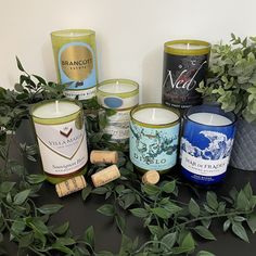 several different types of candles on a table next to some plants and wine corks