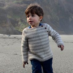 a little boy walking on the beach with his feet in the sand and wearing a sweater