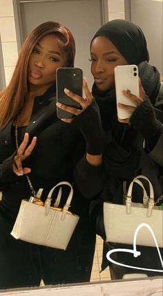 two women taking selfies with their cell phones in front of a mirror and holding purses