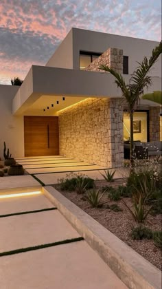 a modern house is lit up at night with lights on the side and palm trees in front