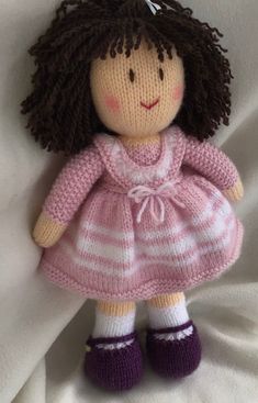 a knitted doll with brown hair wearing a pink dress and purple shoes on a white sheet