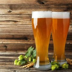 two glasses of beer sitting next to each other on top of a wooden table with hops