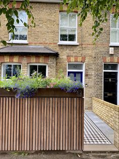 a wooden fence with flowers growing on it in front of a brick building and blue door