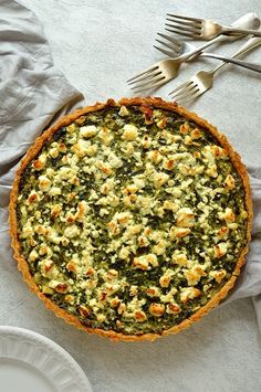 a quiche with spinach, cheese and other toppings sits on a table next to silverware