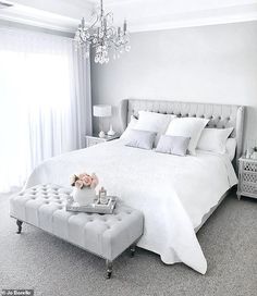 a white bedroom with a chandelier, bed and two tables in front of the window