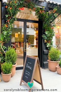 a sign in front of a store with potted plants on the outside and an open door