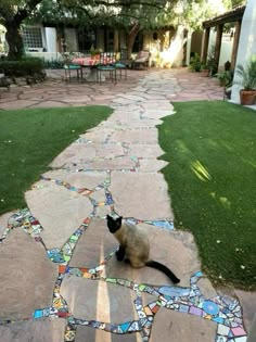 a cat sitting on top of a stone walkway next to a lush green yard and patio