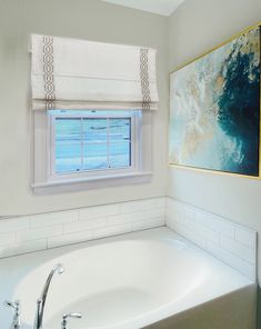 a bath tub sitting under a window next to a painting on the wall in a bathroom