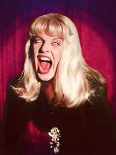 a blonde woman with her mouth open in front of a red curtain and purple background